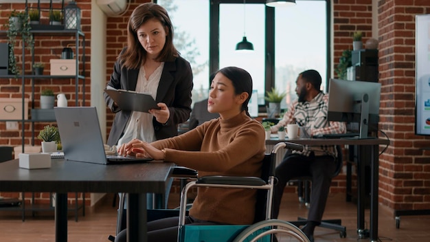 Asian woman in wheelchair brainstorming ideas with colleague, doing teamwork to plan business strategy at startup job. People collaborating in office to work together on development.