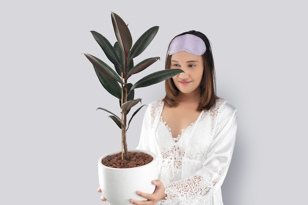 Asian woman in a white satin nightgown wearing lace robe holding rubber plant in white pot.