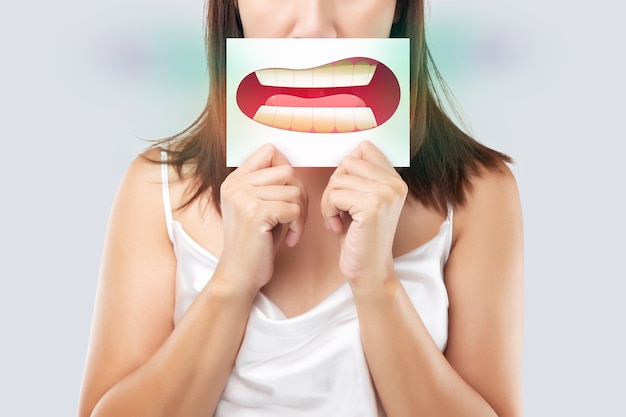Donna asiatica nell'abbigliamento bianco che tiene una carta bianca con l'immagine del fumetto dei denti gialli della sua bocca sullo sfondo grigio, alitosi o alitosi, il concetto con gengive e denti sanitari Foto Premium