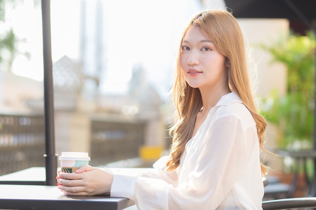 Азиатская женщина в костюме с бронзовыми волосами сидит на стуле в кафе и держит чашку кофе