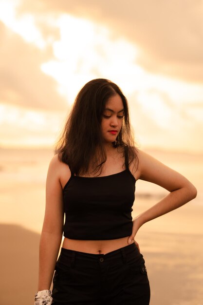 休暇中に平らな表情でビーチの砂の上に立っている黒い服と長い黒髪のアジアの女性