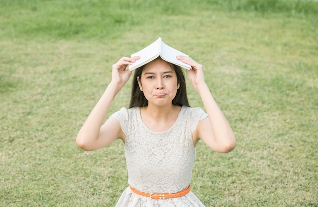 Asian woman with bored emotion with a book on top of her head on blurred grass floor background