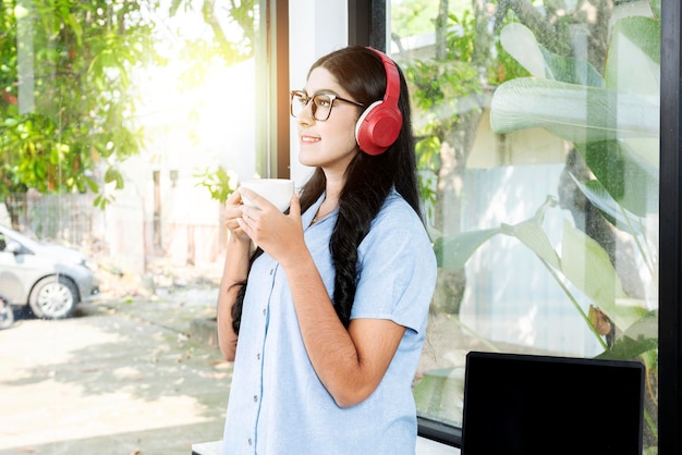 Asian woman with eyeglass listening to music with headphones and holding a cup of coffee with a laptop on the table