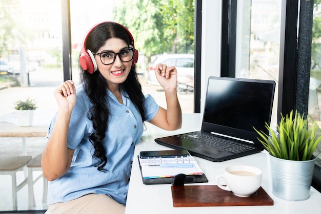 안경을 쓴 아시아 여성, 헤드폰을 끼고 노트북과 노트북, 휴대폰, 탁자 위에 커피 한 잔