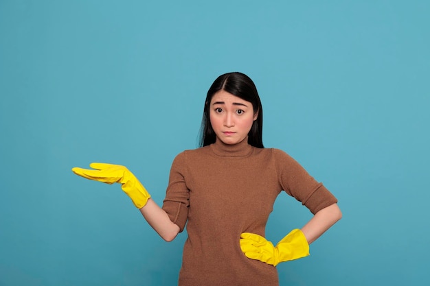 Asian woman with fearful facial expression pointing hand aside\
at copy space and wearing yellow gloves for safety, housewife\
worker, cleaning home concept, stressed worried female
