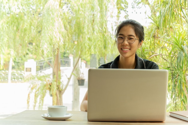 Donna asiatica con gli occhiali che sorride per mezzo del computer portatile con la tazza di caffè sulla tabella.