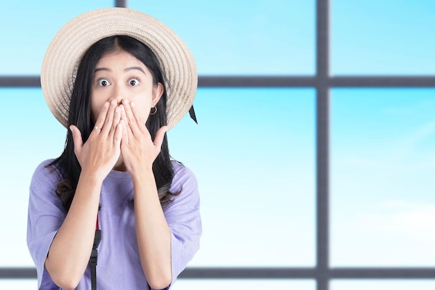 Asian woman with a hat and camera standing with a shocked expression inside the building with blue sky background