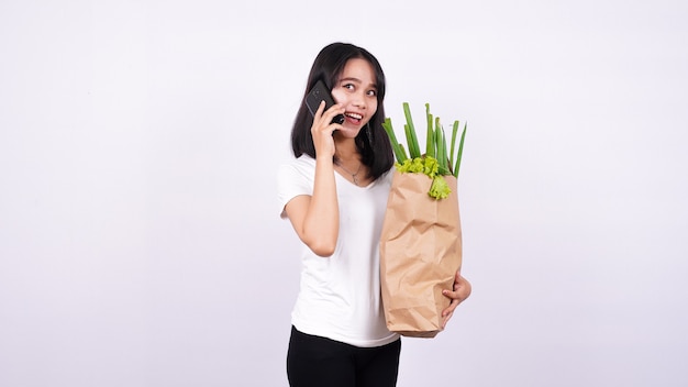 Asian woman with paper bag of fresh vegetables and talking on a phone with isolated white surface