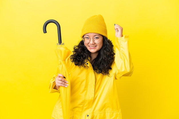 勝利を祝う黄色の背景に分離された防雨コートと傘を持つアジアの女性