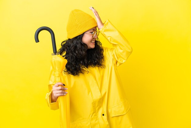 黄色の背景で隔離の防雨コートと傘を持つアジアの女性は何かを実現し、解決策を意図しています
