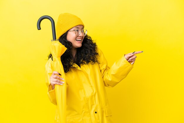 Азиатская женщина с непромокаемым пальто и зонтиком изолирована на желтом фоне, указывая пальцем в сторону и представляет продукт