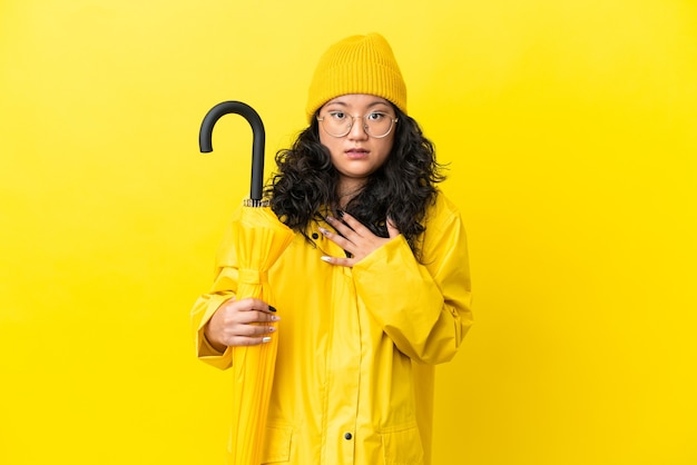 Asian woman with rainproof coat and umbrella isolated on yellow background surprised and shocked while looking right