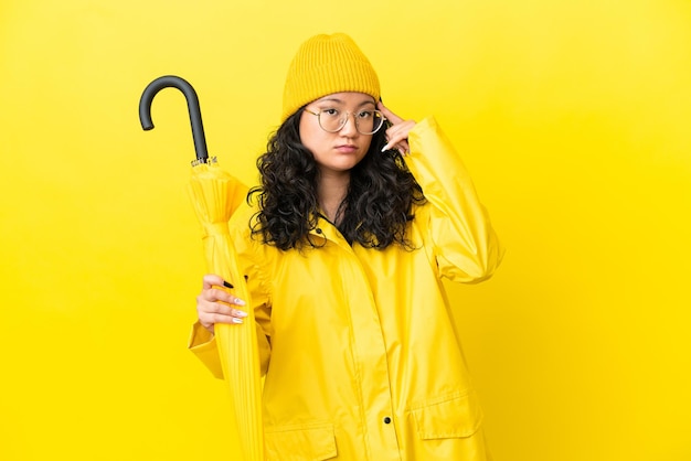 Asian woman with rainproof coat and umbrella isolated on yellow background thinking an idea