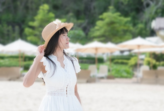 Asian woman with sea sand concept. lady standing with the wind\
blows my hair on blur image of white umbrella at sea sand\
beach