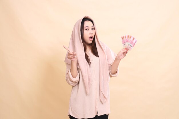 Asian woman with shocked expression at the camera pointing her hand to the right and carrying rupiah