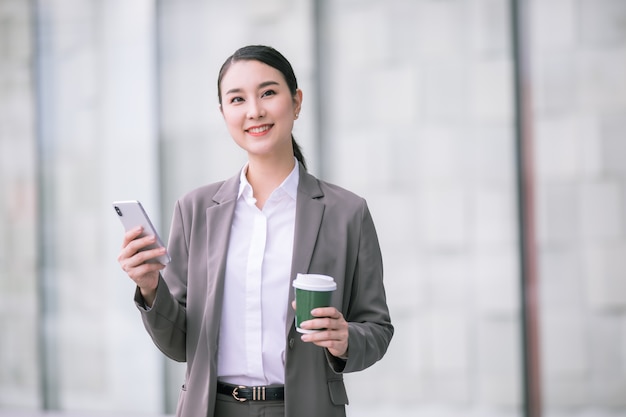 Asian woman with smartphone standing against street blurred building. Fashion business photo of beautiful girl in casual suite with phone and cup of coffee.
