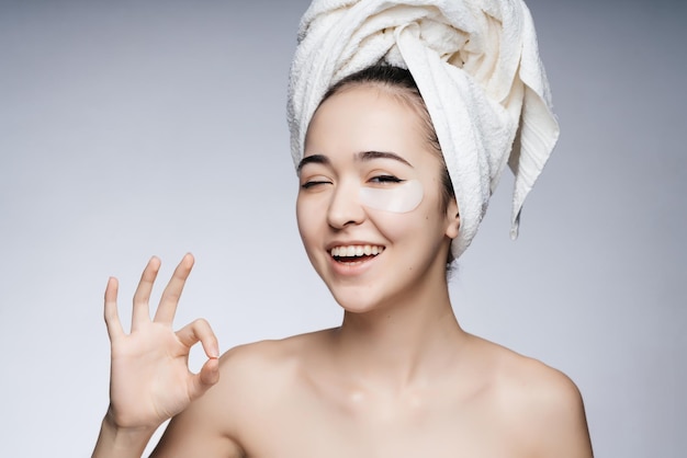 Asian woman with a towel on her head and bare shoulders pasted a patch under her eyes and smiling shows her hand an OK sign