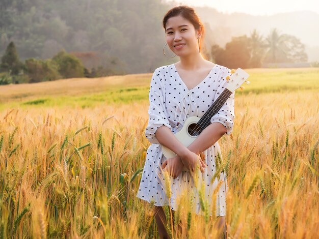 Asian woman with ukulele in barley field at sunset time.