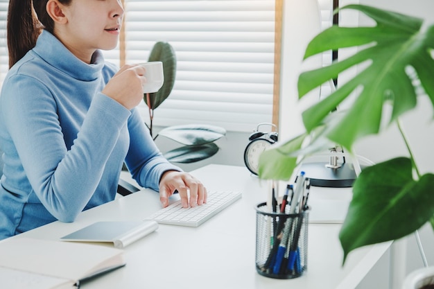 Asian woman working from home using computer and drinking coffee in her bedroom document finance and conference online meeting for new projects