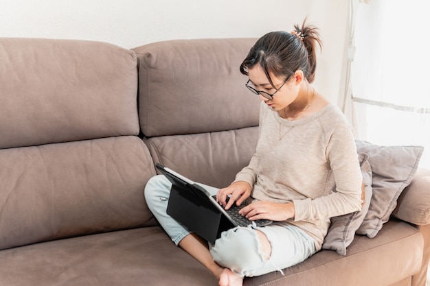 写真 ソファでノートパソコンやタブレットを使って在宅勤務するアジア人女性