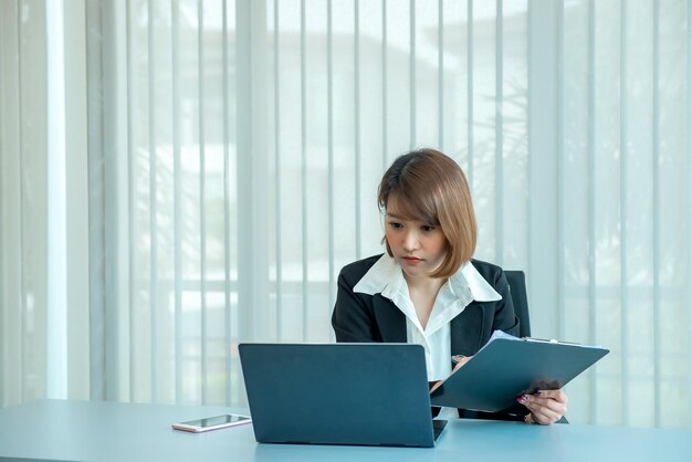 オフィスで残業しているアジアの女性ビジネスマンたくさんの仕事ラップトップでファイルをチェックタイの人々はオフィスの制服を着ています