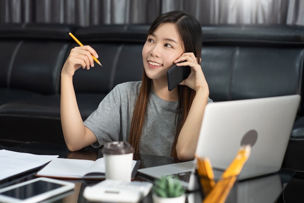 Азиатская женщина работает с ноутбуком и документом, говоря смартфон в помещении гостиной