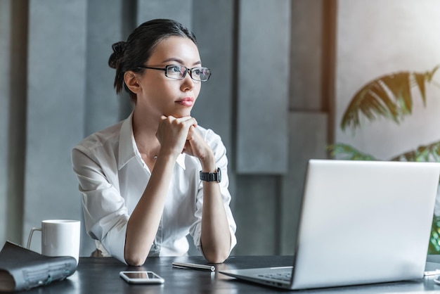 Donna asiatica che lavora con il computer portatile e pensa vicino al posto di lavoro