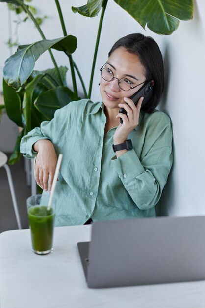 Asian woman works online has phone conversation with client\
works on modern laptop computer drinks fresh healthy smoothie poses\
in home office wears spectacles and shirt looks thoughtfully\
away