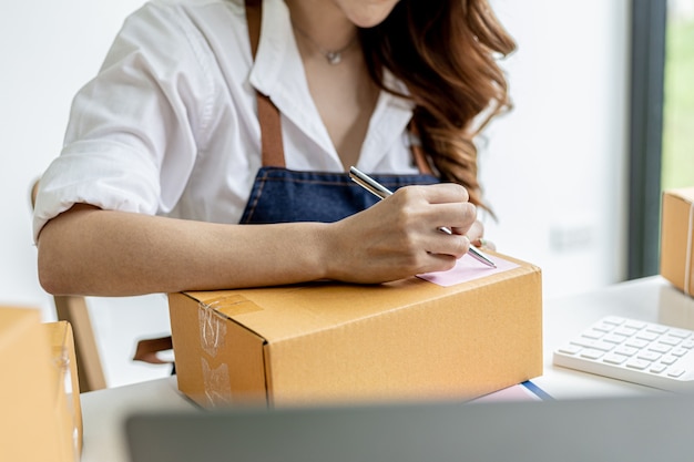 小包箱に顧客の配送情報を書いているアジアの女性は、オンラインストアを所有しており、民間の宅配会社を通じて顧客に商品を配送しています。オンライン販売のコンセプト。
