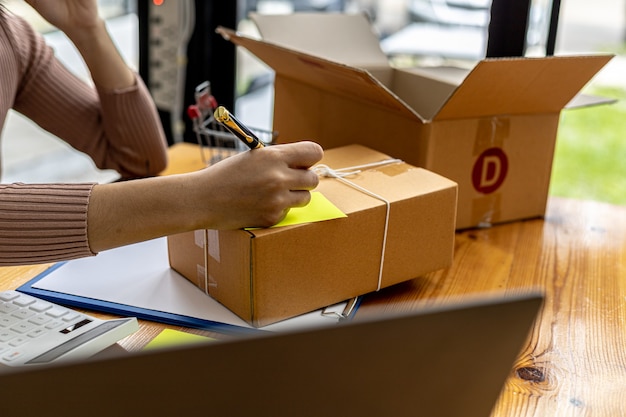 Asian woman writing customer's shipping information on parcel box, she owns an online store, she ships products to customers through a private courier company. Online selling concept.