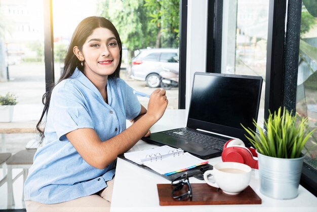 Asian woman writing something on a notebook with a laptop and a cup of coffee on the table