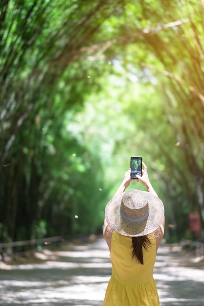 黄色のドレスと帽子をかぶったアジア人女性緑の竹トンネルで旅行タイのナコンナーヨックにあるチュラブホーンワナラム寺院で携帯電話で写真を撮る幸せな旅行者