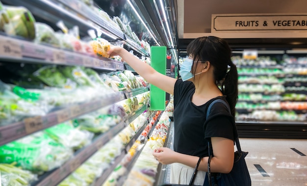 アジアの女性は食料品店で買い物をし、バスケットを持って、健康マスクを着用しています