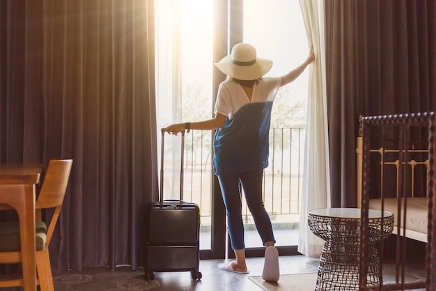 写真 アジアの女性は荷物を持ってホテルの部屋に滞在しています部屋のカーテンとドアを開けて外の景色を眺める休日の旅行のコンセプトヴィンテージトーン