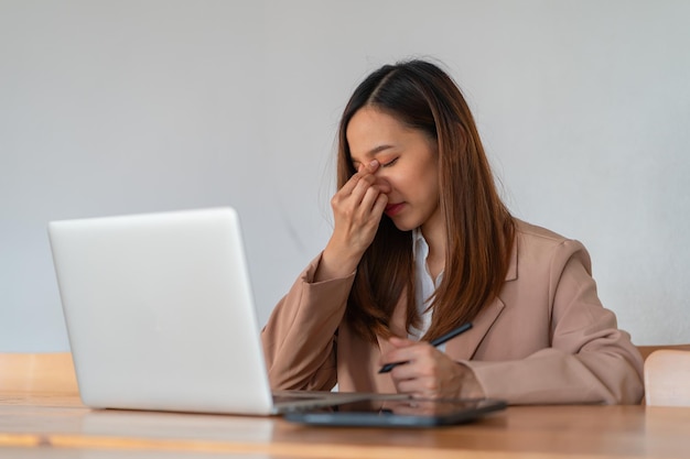 アジアの女性はノートパソコンで働くときにストレスを受けています オフィスで頭痛を感じる疲れたアジアのビジネス女性は 仕事で病気を感じてコピースペース