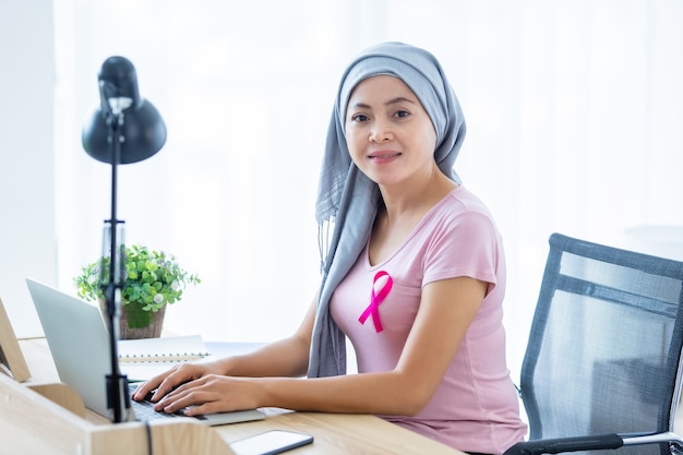 A asian women disease mammary cancer patient with pink ribbon wearing headscarf After treatment to chemotherapy with working business at laptop in office at home,medicine concept