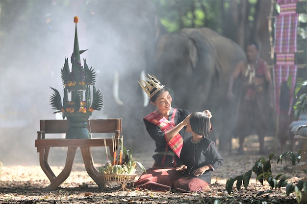 Asian women in native traditional costume sitting by harvest ceremony set on table against elephants.