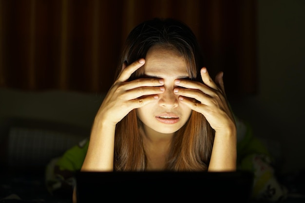写真 アジアの女性は、暗い場所でコンピューターを見てオンラインで映画を見ることによる眼精疲労に苦しんでいます