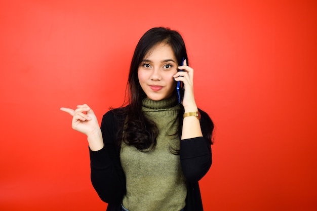 asian women using black sweater looking and pointing aside talking on phone isolated on red