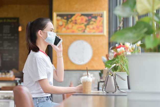 アジアの女性はサージカルマスクを着用し、カフェでスマートフォンを使用しています