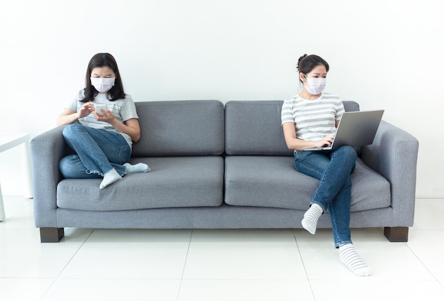 Asian women wearing masks working at home using notebook and smartphone to reduce spreading of coronavirus infection during crisis of Covid-19. Work from home and social distancing concept.