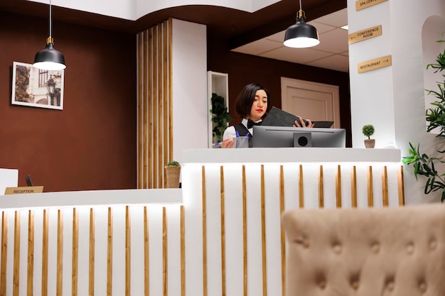 Photo asian worker checking booking enquiries at reception desk, preparing to welcome guests upon arrival. receptionist working at hotel counter in lobby looking at registration forms, accommodation.