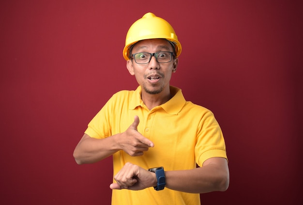 Asian worker man wearing helmet  in hurry pointing to watch time upset and angry for deadline delay