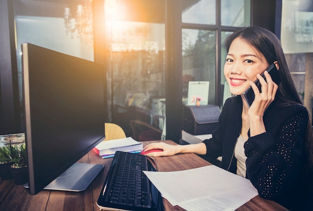 ホームオフィスのコンピューターを使用して、幸せそうな顔で携帯電話で話しているアジアの働く女性