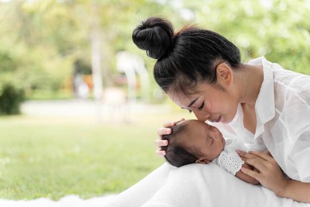 新生児を抱いたアジアの若い美しい母親が眠っていて、愛情を込めて感じ、優しく触れて、公園の緑の芝生に座っています