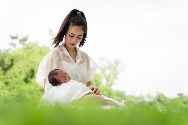 Asian young beautiful mother holding her newborn is sleeping and feel with love and touching gently then sitting on green grass in the park