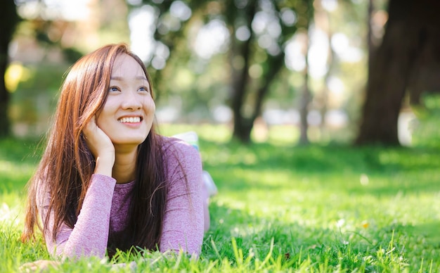 Asian young beautiful woman wearing sports shirt smiling lying on green grass on the background of a green summer park.