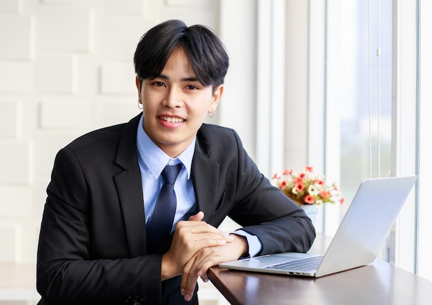 写真 アジアの青年実業家は、朝、オフィスの窓の近くのラップトップでキーボードを入力しながら座っている黒いスーツを着てスマートでハンサムです。