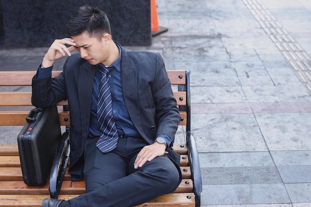 Asian young businessman stress sitting on bench in front office with his hands touching his head To