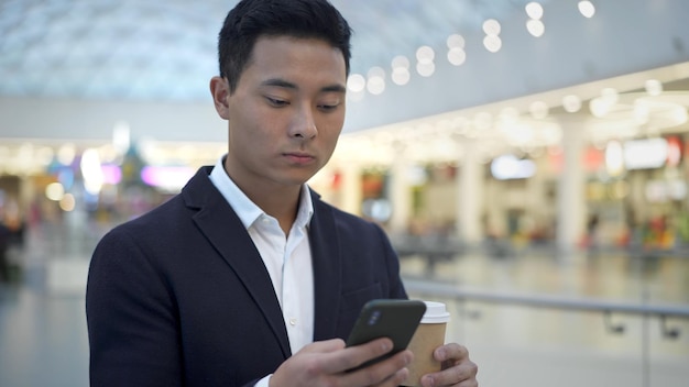 Азиатский молодой бизнесмен в костюме пьет кофе и пишет смс по телефону